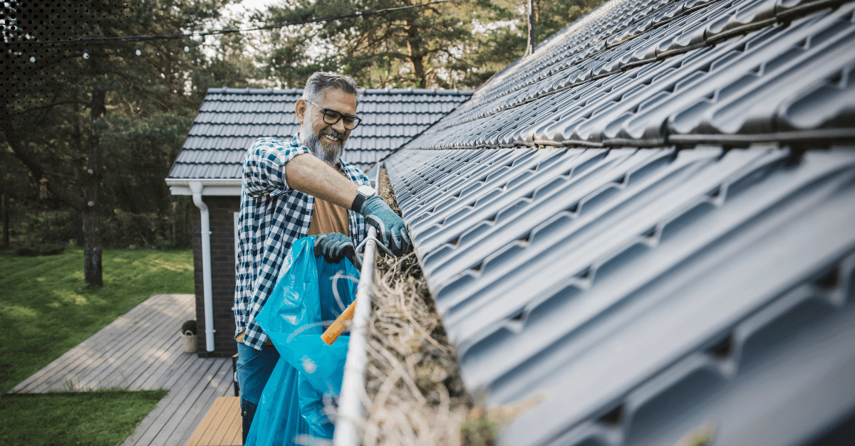 A cheerful man wearing gloves and a plaid shirt cleans debris from a gutter attached to a modern metal roof, placing the waste into a blue bag.