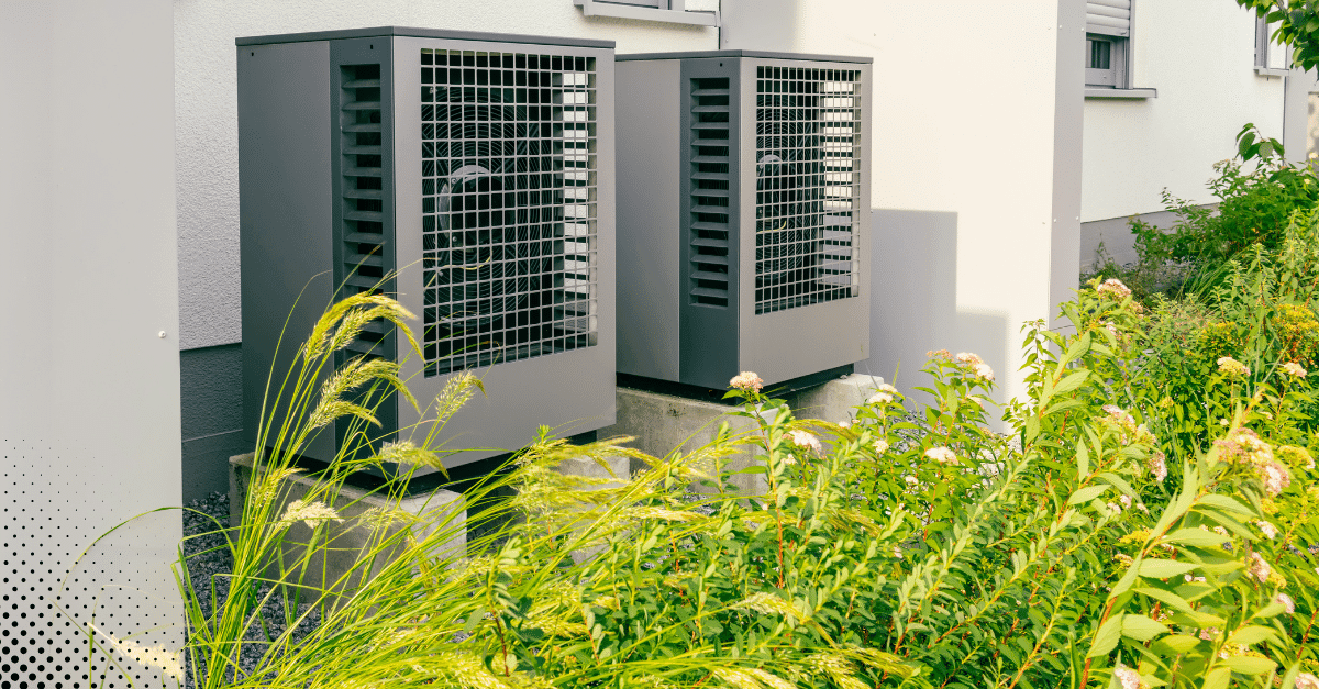 Two outdoor HVAC units beside a house surrounded by landscaping and greenery.